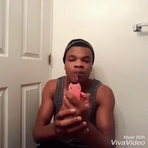 A Man In A Tank Top Is Holding A Pink Phone In Front Of A White Door Made With Vivavideo GIF