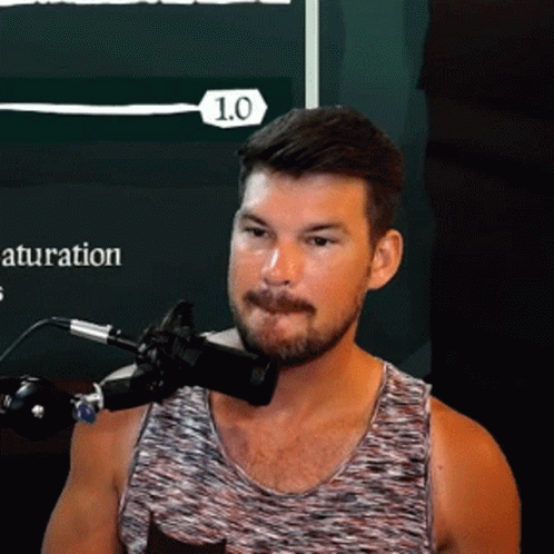 A Man In A Tank Top Is Speaking Into A Microphone With The Number 1.0 On The Bottom Right GIF