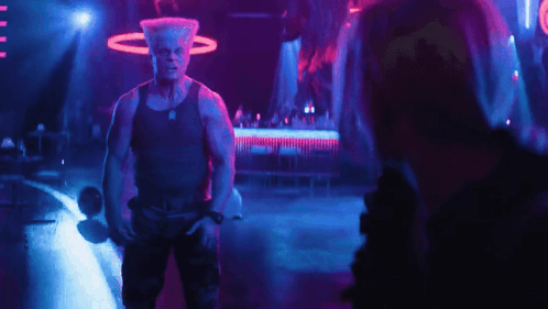 A Man In A Tank Top Stands In A Dark Room GIF