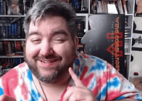 A Man In A Tie Dye Shirt Is Smiling In Front Of A Wing Commander Sign GIF