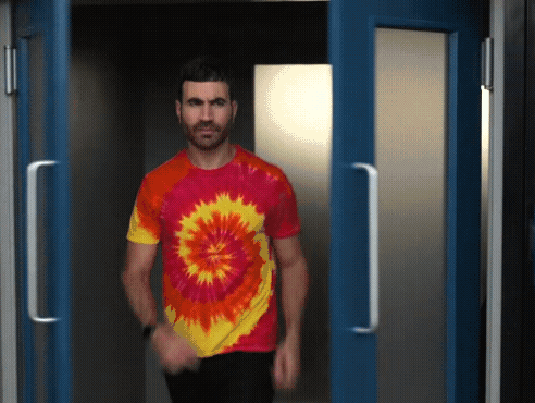 A Man In A Tie Dye Shirt Is Walking Through A Doorway GIF