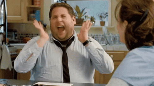 A Man In A Tie Is Laughing In A Kitchen While Sitting At A Table With A Woman . GIF