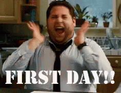A Man In A Tie Is Sitting At A Table With His Hands In The Air And The Words First Day Written Below Him . GIF