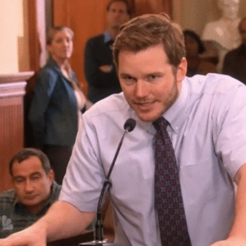 A Man In A Tie Is Speaking Into A Microphone In Front Of A Crowd GIF