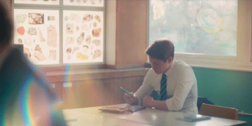 A Man In A Tie Sits At A Desk In A Classroom Looking At His Phone GIF