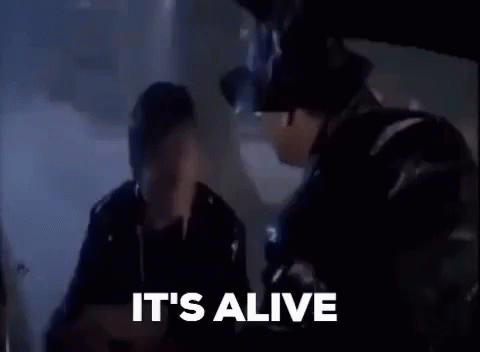 A Man In A Top Hat Is Talking To Another Man In A Dark Room And The Words It 'S Alive Are Visible . GIF