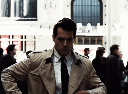 A Man In A Trench Coat And Tie Stands In A Crowd GIF