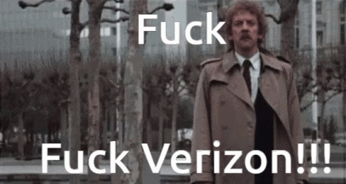 A Man In A Trench Coat And Tie Stands In Front Of Trees And Says Fuck Verizon !!! GIF