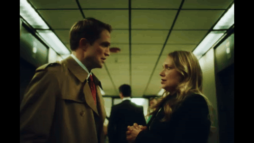 A Man In A Trench Coat And Tie Talks To A Woman GIF