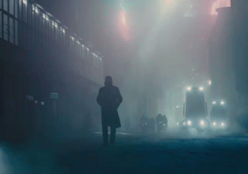 A Man In A Trench Coat Stands In A Foggy City Street GIF