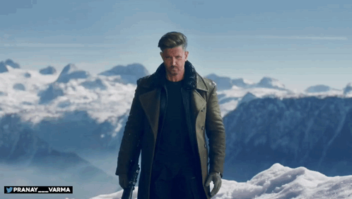 A Man In A Trench Coat Stands On Top Of A Snow Covered Mountain With The Name Pranay Varma Below Him GIF