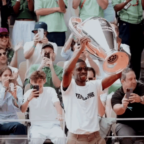 A Man In A Triumphe Shirt Holds Up A Trophy In Front Of A Crowd Of People GIF