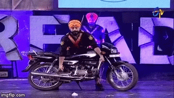 A Man In A Turban Is Standing On A Motorcycle In Front Of A Sign That Says " Real " GIF