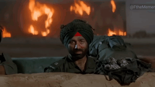 A Man In A Turban Looks Out From Behind A Wall With A Fire In The Background GIF