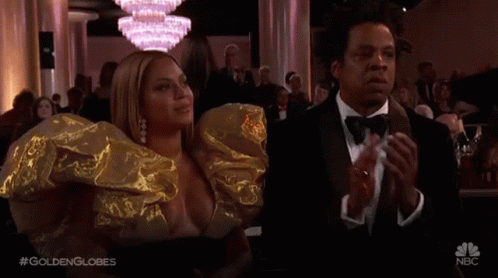 A Man In A Tuxedo And A Woman In A Gold Dress Are Applauding At A Golden Globes Event . GIF