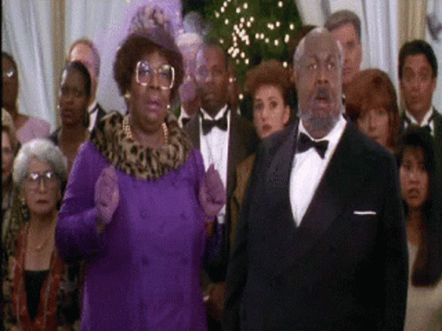 A Man In A Tuxedo And A Woman In A Purple Coat Stand In Front Of A Crowd Of People GIF