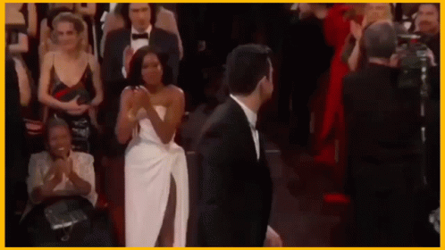 A Man In A Tuxedo And A Woman In A White Dress Are Standing Next To Each Other In Front Of A Crowd . GIF