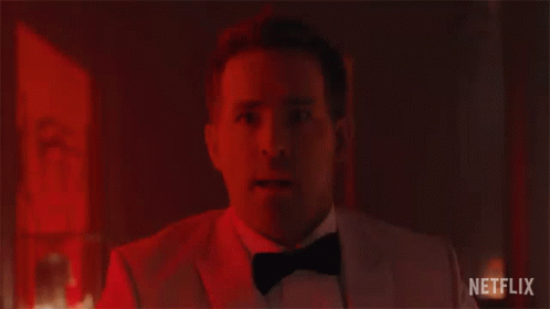 A Man In A Tuxedo And Bow Tie Is Looking At The Camera In A Dark Room . GIF