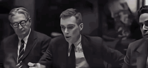 A Man In A Tuxedo And Bow Tie Is Sitting At A Table In A Black And White Photo . GIF
