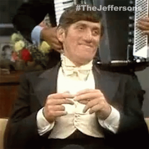 A Man In A Tuxedo And Bow Tie Is Sitting In A Chair And Holding A Piece Of Paper . GIF