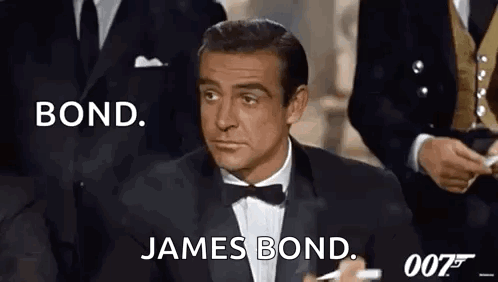 A Man In A Tuxedo And Bow Tie Is Sitting In Front Of A Group Of Men In Suits . GIF