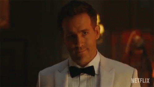 A Man In A Tuxedo And Bow Tie Is Smiling In A Dark Room . GIF
