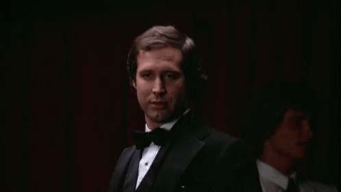 A Man In A Tuxedo And Bow Tie Is Smoking A Cigarette In A Dark Room . GIF