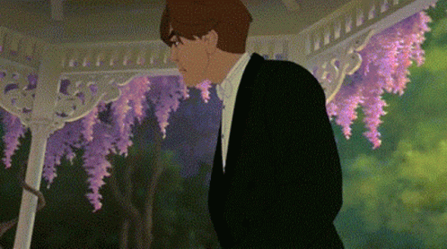 A Man In A Tuxedo And Bow Tie Is Standing In Front Of A Gazebo With Purple Flowers . GIF
