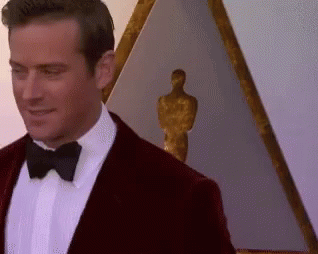 A Man In A Tuxedo And Bow Tie Is Standing In Front Of A Statue Of An Oscar Winner . GIF