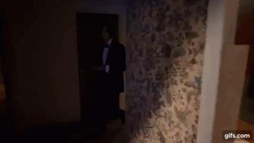 A Man In A Tuxedo And Bow Tie Is Walking Through A Doorway . GIF