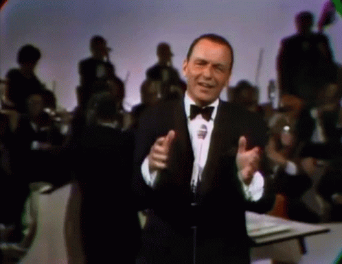 A Man In A Tuxedo And Bow Tie Singing Into A Microphone GIF