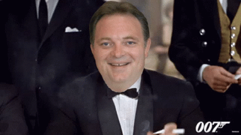 A Man In A Tuxedo And Bow Tie Smoking A Cigarette With The Number 007 Behind Him GIF