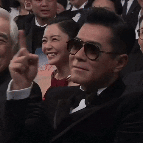 A Man In A Tuxedo And Sunglasses Is Sitting In A Crowd GIF