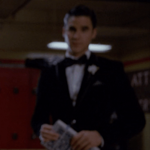 A Man In A Tuxedo Holds A Piece Of Paper In Front Of A Sign That Says Cafe GIF