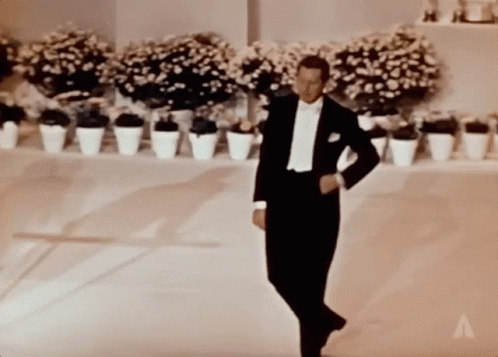 A Man In A Tuxedo Is Dancing In Front Of Potted Flowers GIF