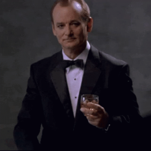 A Man In A Tuxedo Is Holding A Glass Of Whiskey And Pointing At The Camera With The Words You Da Man Above Him GIF