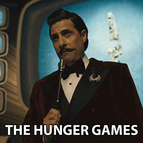 A Man In A Tuxedo Is Holding A Microphone And The Hunger Games Is Written Below Him GIF