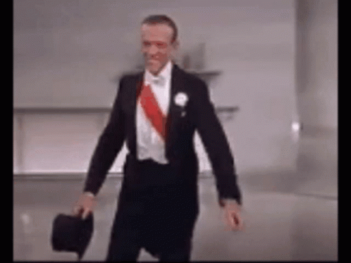 A Man In A Tuxedo Is Holding A Top Hat And Walking . GIF