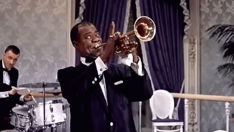 A Man In A Tuxedo Is Playing A Trumpet In A Room With A Drummer In The Background . GIF