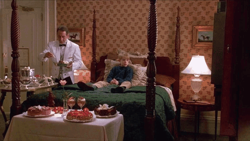 A Man In A Tuxedo Is Serving A Dessert To A Young Boy Laying On A Bed GIF