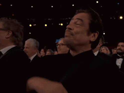 A Man In A Tuxedo Is Sitting In A Dark Room With Other People GIF