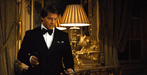 A Man In A Tuxedo Is Standing In A Room With A Lamp GIF