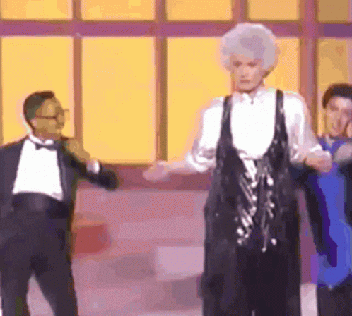 A Man In A Tuxedo Is Standing Next To A Woman In A Black Dress Who Is Dancing . GIF
