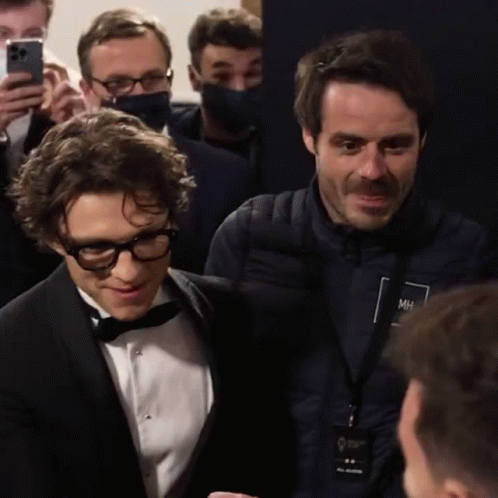 A Man In A Tuxedo Is Talking To Another Man In A Jacket With Mh On It GIF
