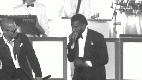 A Man In A Tuxedo Sings Into A Microphone While Another Man Stands Behind Him Playing A Keyboard GIF