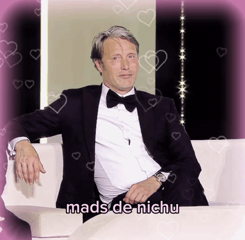 A Man In A Tuxedo Sits On A White Couch With The Words Mads De Nichu Written Below Him GIF