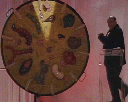 A Man In A Tuxedo Stands In Front Of A Large Circular Display Of Various Food Items GIF