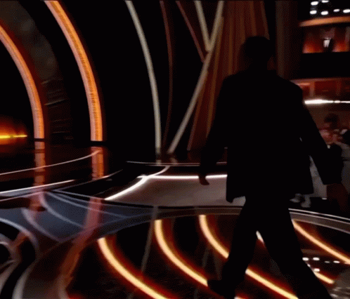 A Man In A Tuxedo Stands On A Stage With A Reflection Of Himself GIF