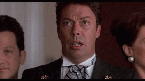 A Man In A Tuxedo With A Bow Tie Looks At The Camera With A Woman Behind Him GIF
