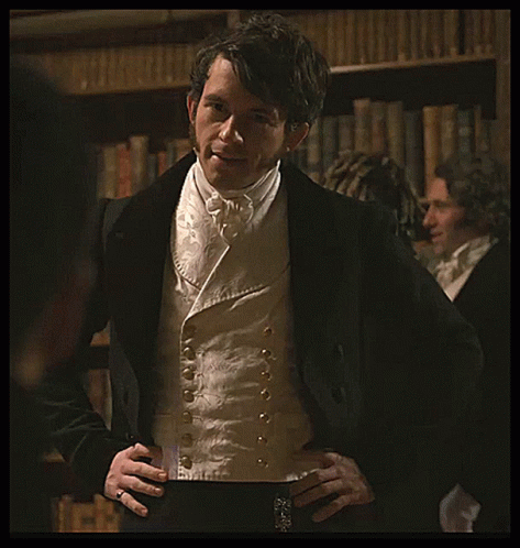 A Man In A Tuxedo With His Hands On His Hips Stands In Front Of A Bookshelf GIF
